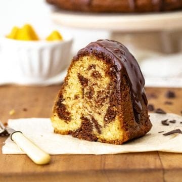 A single slice of marble chocolate orange bundt cake on white paper on wooden table, a fork, white bowl and cake stand in background.