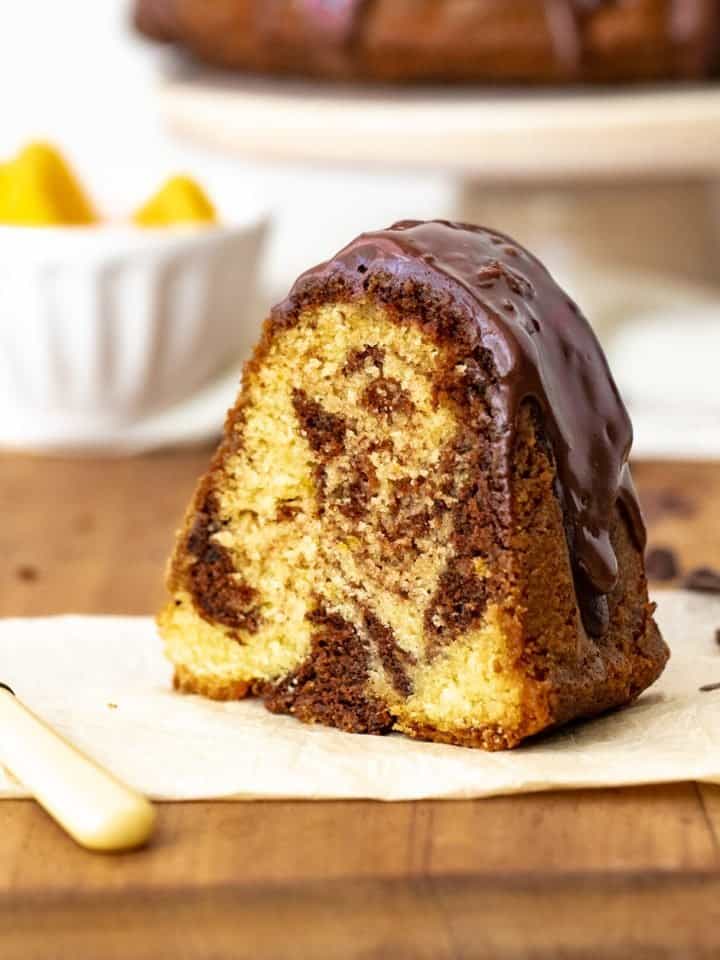 A single slice of marble chocolate orange bundt cake on white paper on wooden table, a fork, white bowl and cake stand in background.