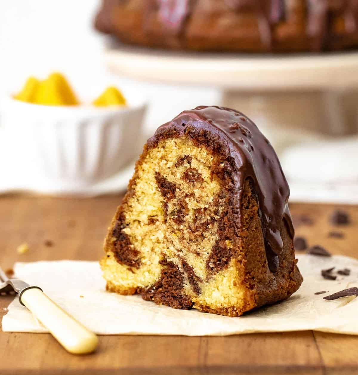 Single slice of marble chocolate orange bundt cake on white paper on wooden table, a fork, white bowl and cake stand in background.