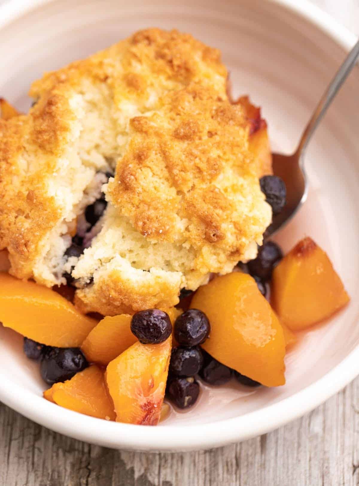 Close up of peach blueberry cobbler serving in white bowl with a spoon.