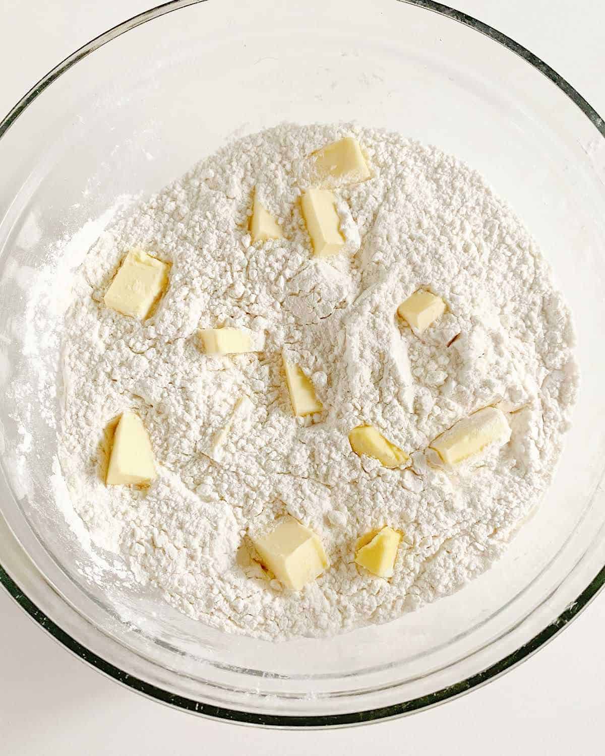 Butter pieces added to flour mixture in a glass bowl on a white surface. 