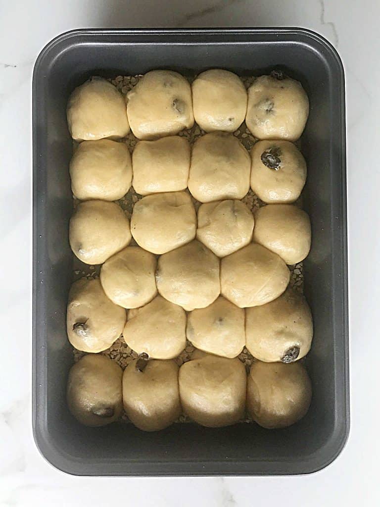 Proofed small raisin buns in a metal rectangular baking pan on a white surface.