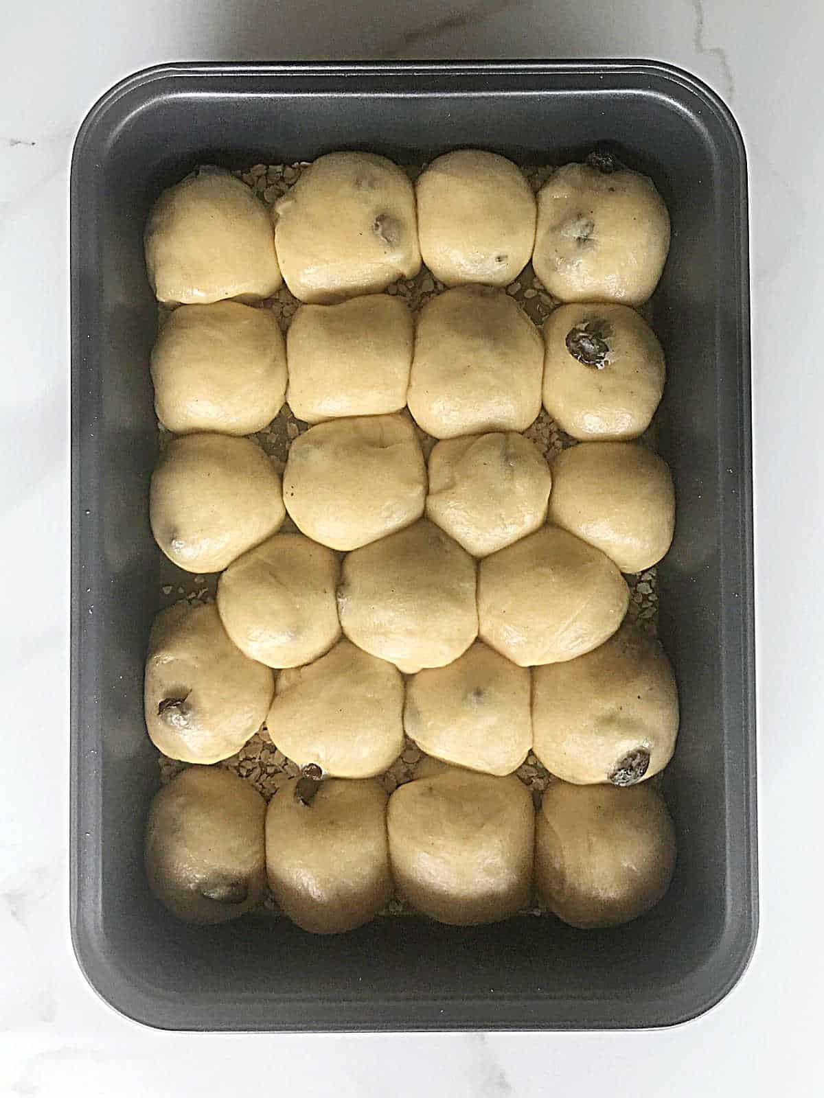Proofed small raisin buns in a metal rectangular baking pan on a white surface.