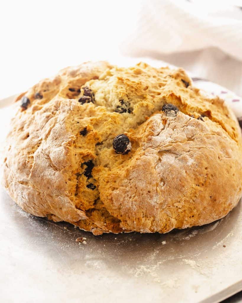 Whole loaf of soda bread with raisins on a metal sheet, white background.