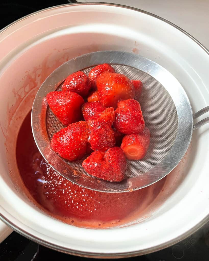 Skimming whole cooked strawberries from a white saucepan.