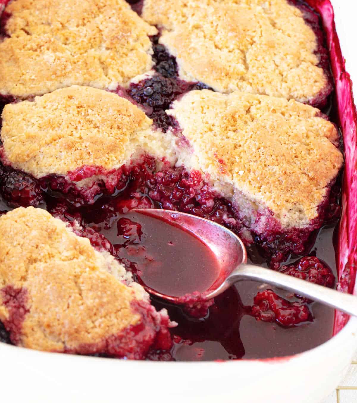 Close up of baked blackberry cobbler with silver spoon in white ceramic dish.