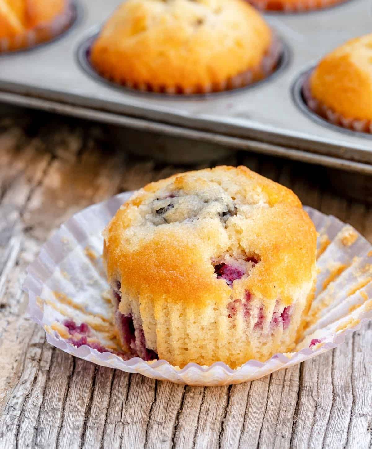 Whole blackberry muffin in opened paper liner on greyish wooden surface; pan of muffins in background.