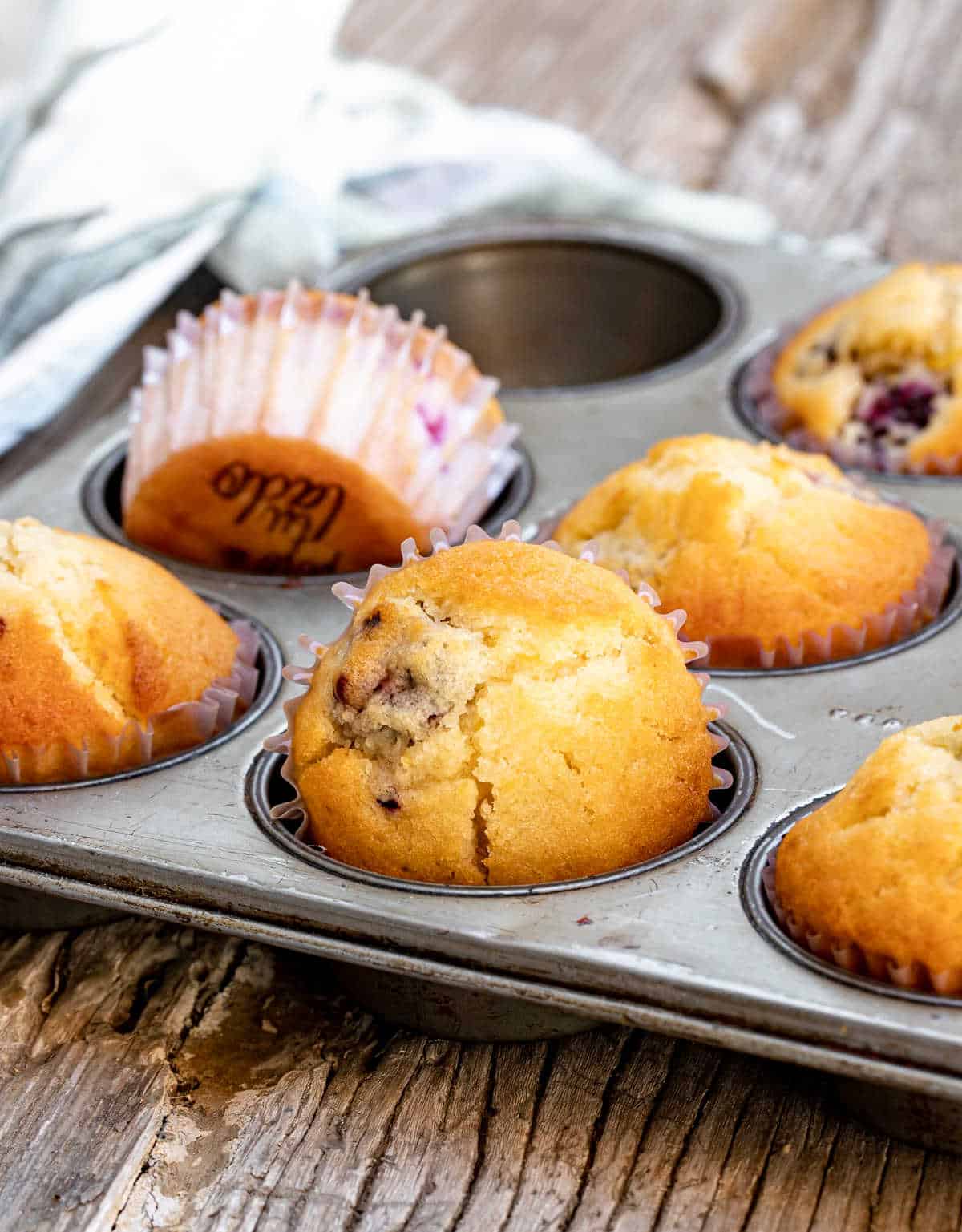 Metal muffin tin with baked blackberry muffins, grey wooden surface, kitchen towel in background.