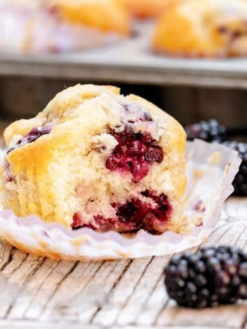 Opened paper liner with half blackberry on top on wooden grey surface, loose blackberries, muffin pan in background.