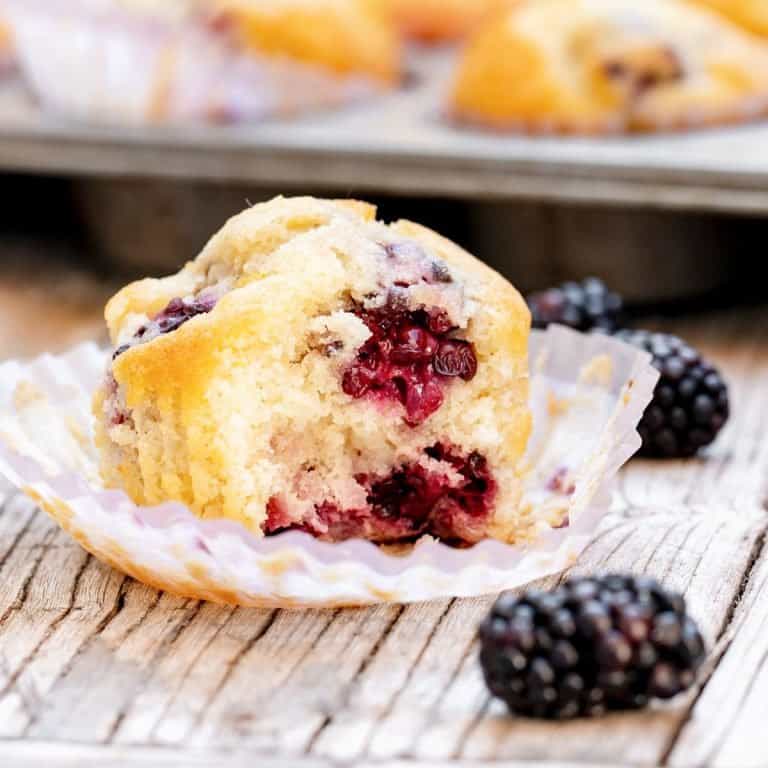 Opened paper liner with half blackberry on top on wooden grey surface, loose blackberries, muffin pan in background.