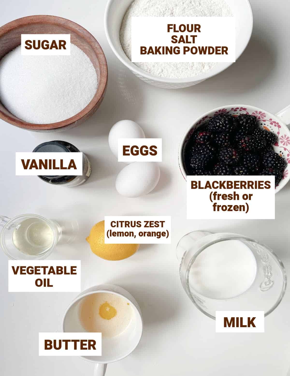 Bowls on white surface with ingredients for blackberry muffins including sugar, butter, oil, milk, eggs, flour mixture.