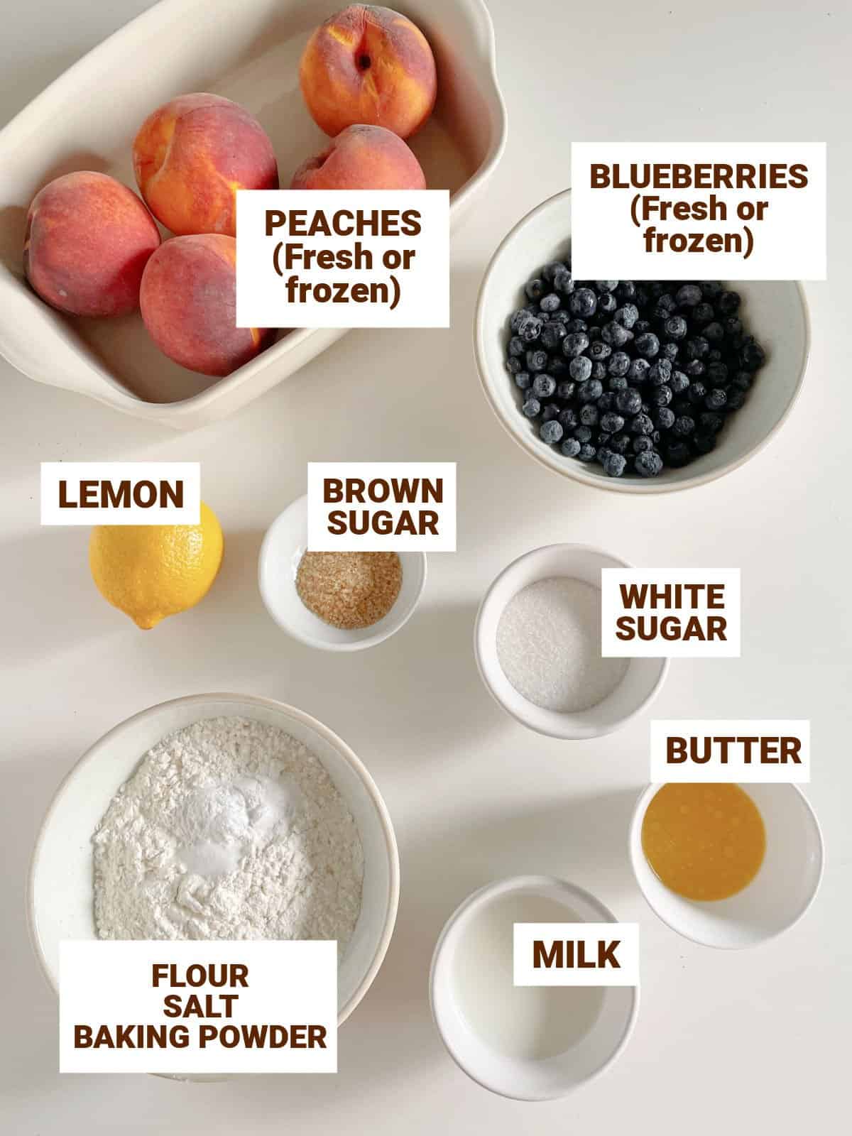 White surface with bowls containing ingredients for blueberry peach cobbler including butter, flour, lemon, brown sugar, milk, fresh fruit.