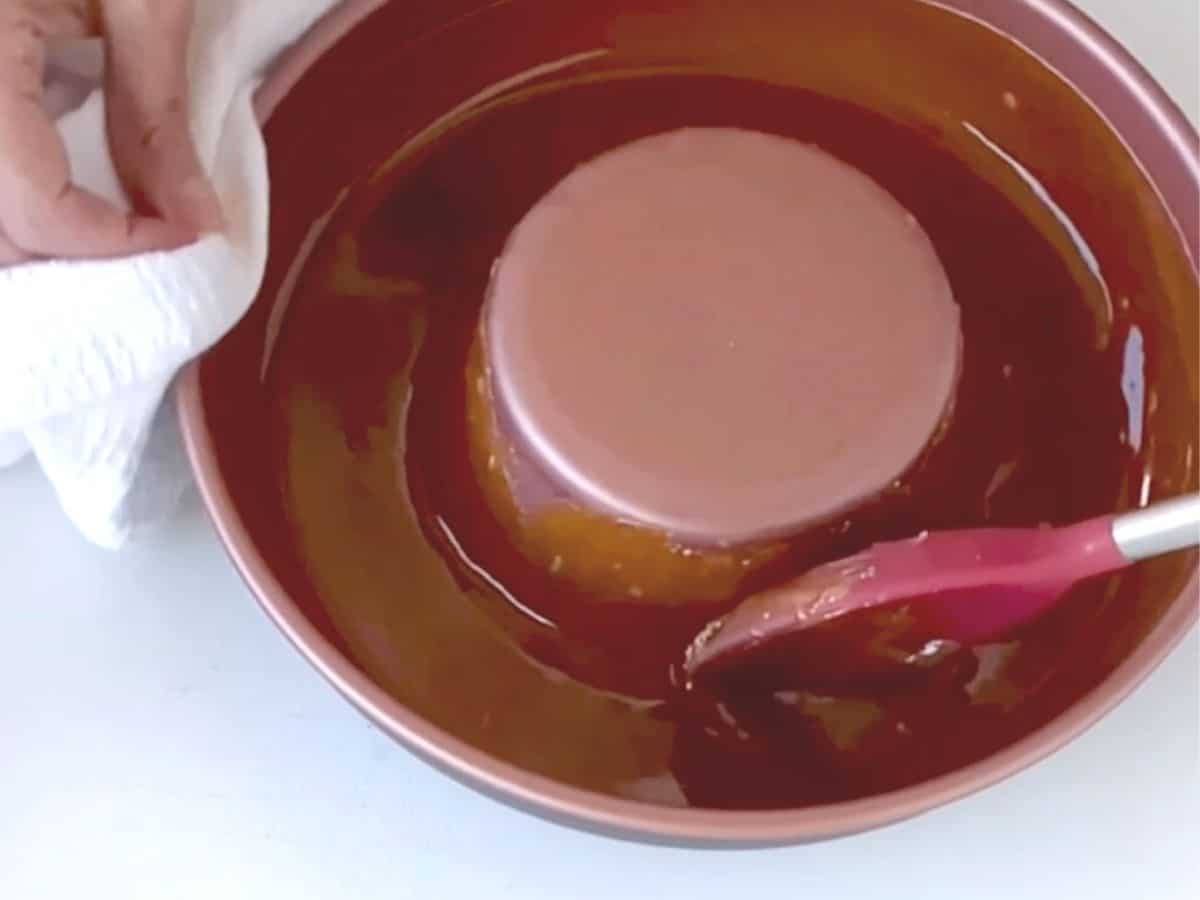 Spreading caramel in flan pan with red silicone spoon on white table.