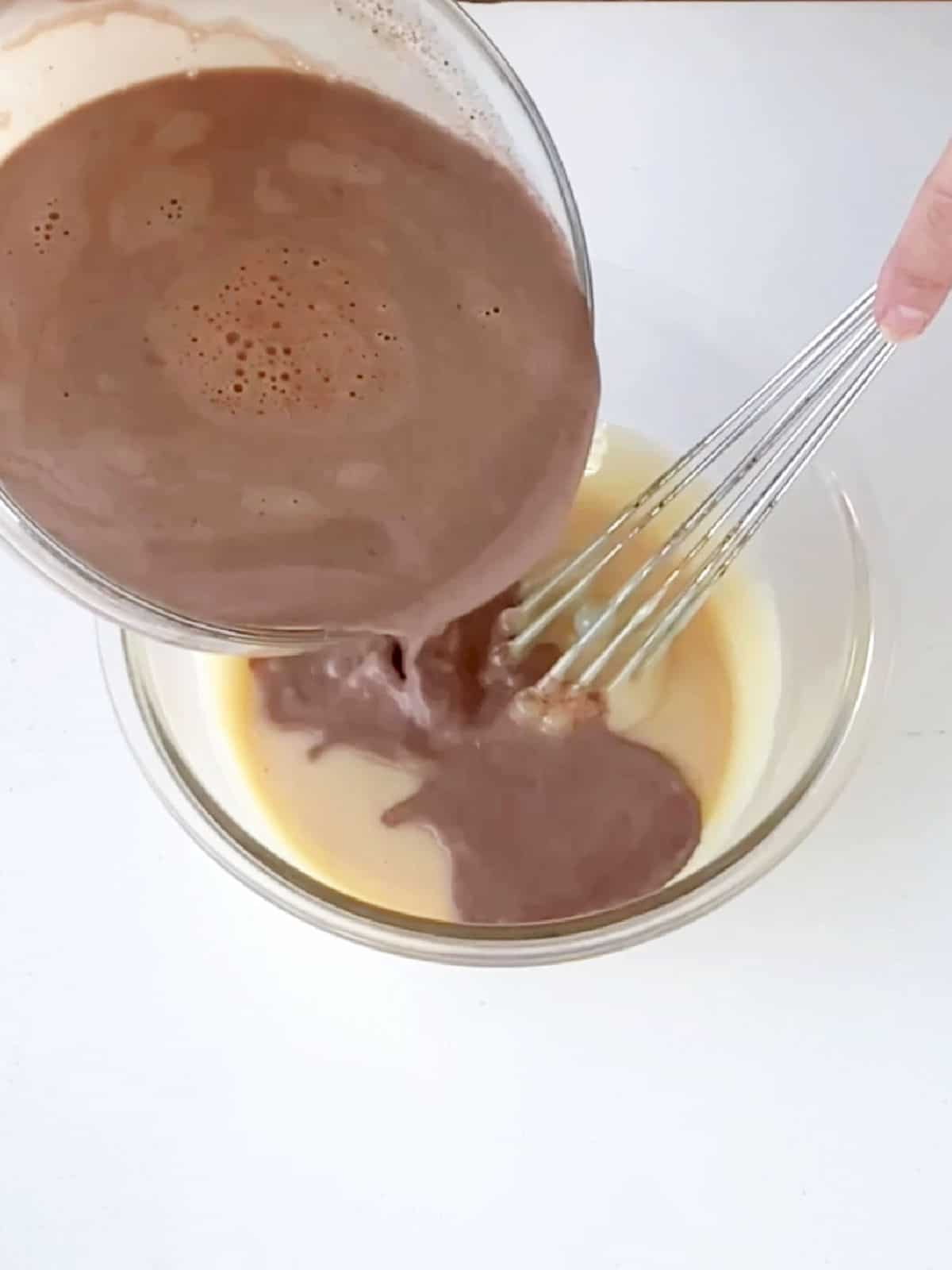 Adding chocolate custard to condensed milk in a glass bowl on a white surface. A hand with a whisk.