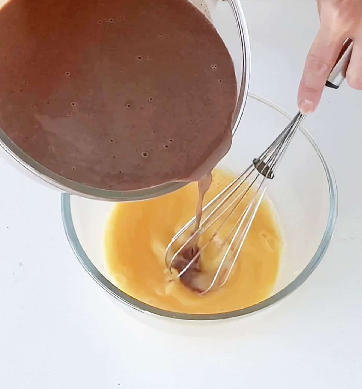 A chocolate custard being poured over beaten eggs in a glass bowl on a white surface. A hand with a whisk.
