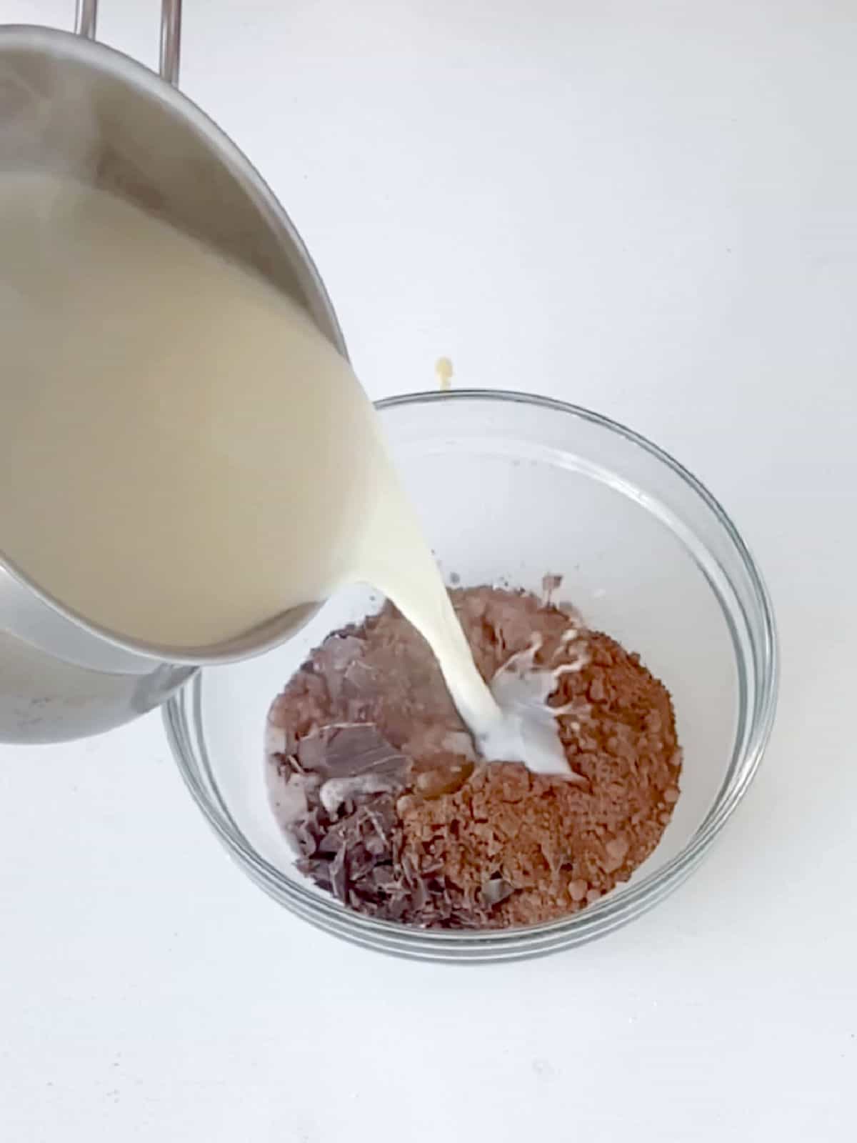 Milk being added to chopped chocolate and cocoa powder in a glass bowl on a white surface. 
