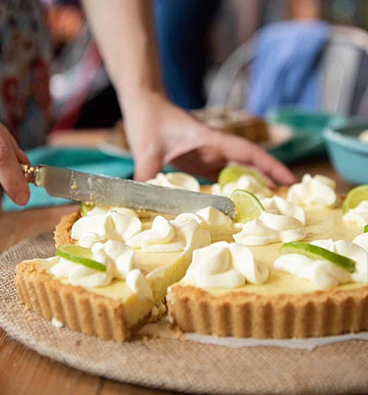 Hands with silver knife cutting a lime pie with cream on top, burlap surface.