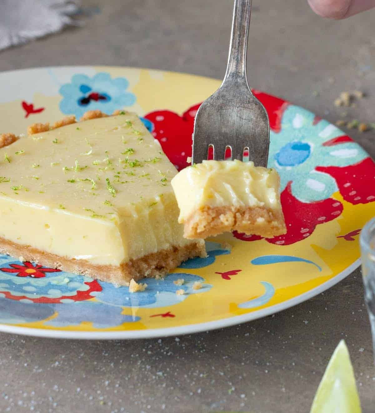 Fork with piece of key lime pie, rest of slice on colorful plate, grey surface.