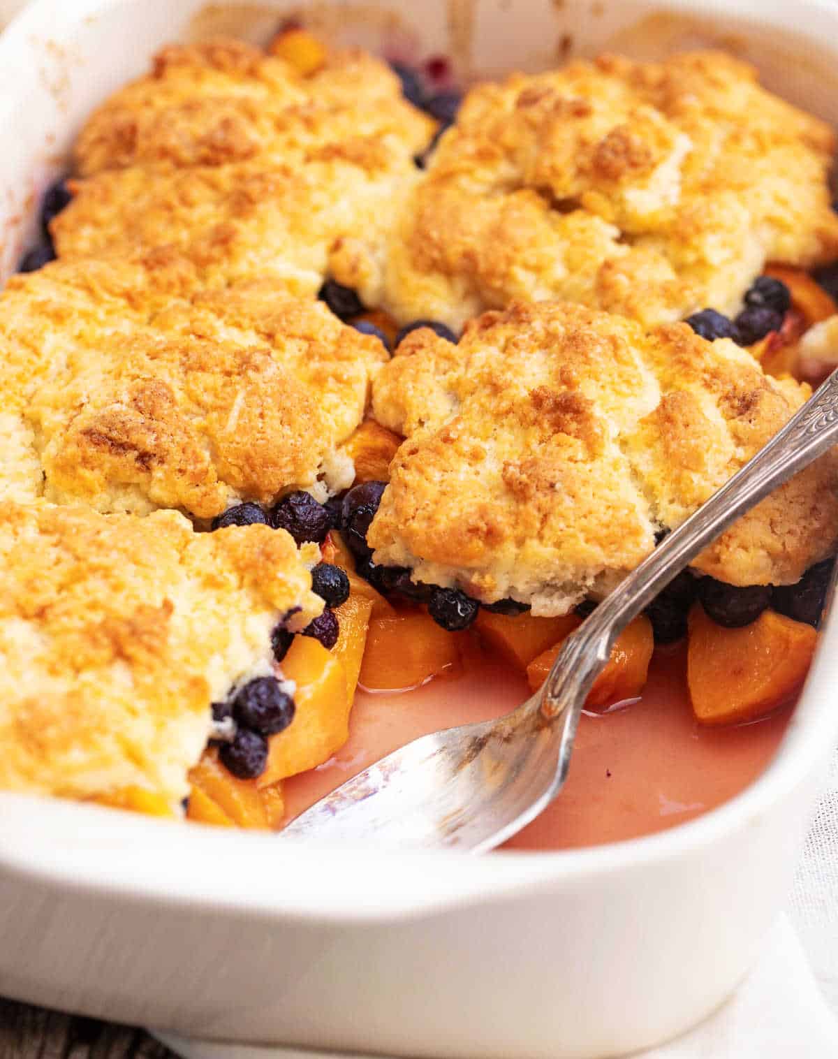 Off white dish with blueberry peach cobbler missing a serving, silver spoon inside.