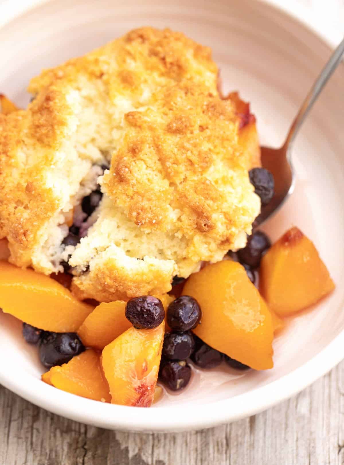 Close up of peach blueberry cobbler serving in white bowl with a spoon.