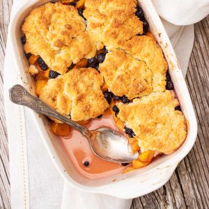 Overview of white dish with silver spoon containing blueberry peach cobbler on white linen and wooden table.