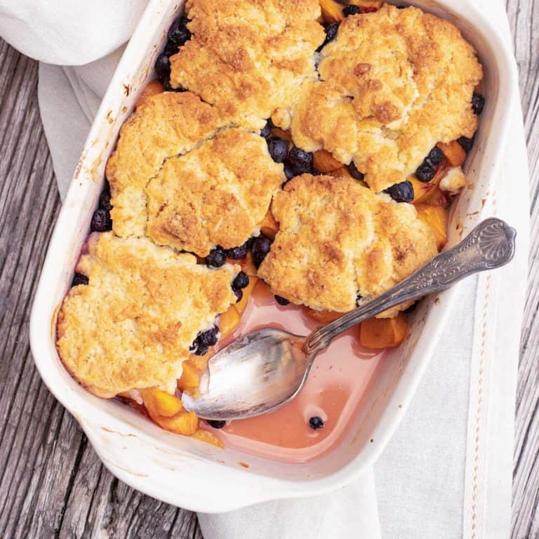 Overview of white dish with silver spoon containing blueberry peach cobbler on white linen and wooden table.