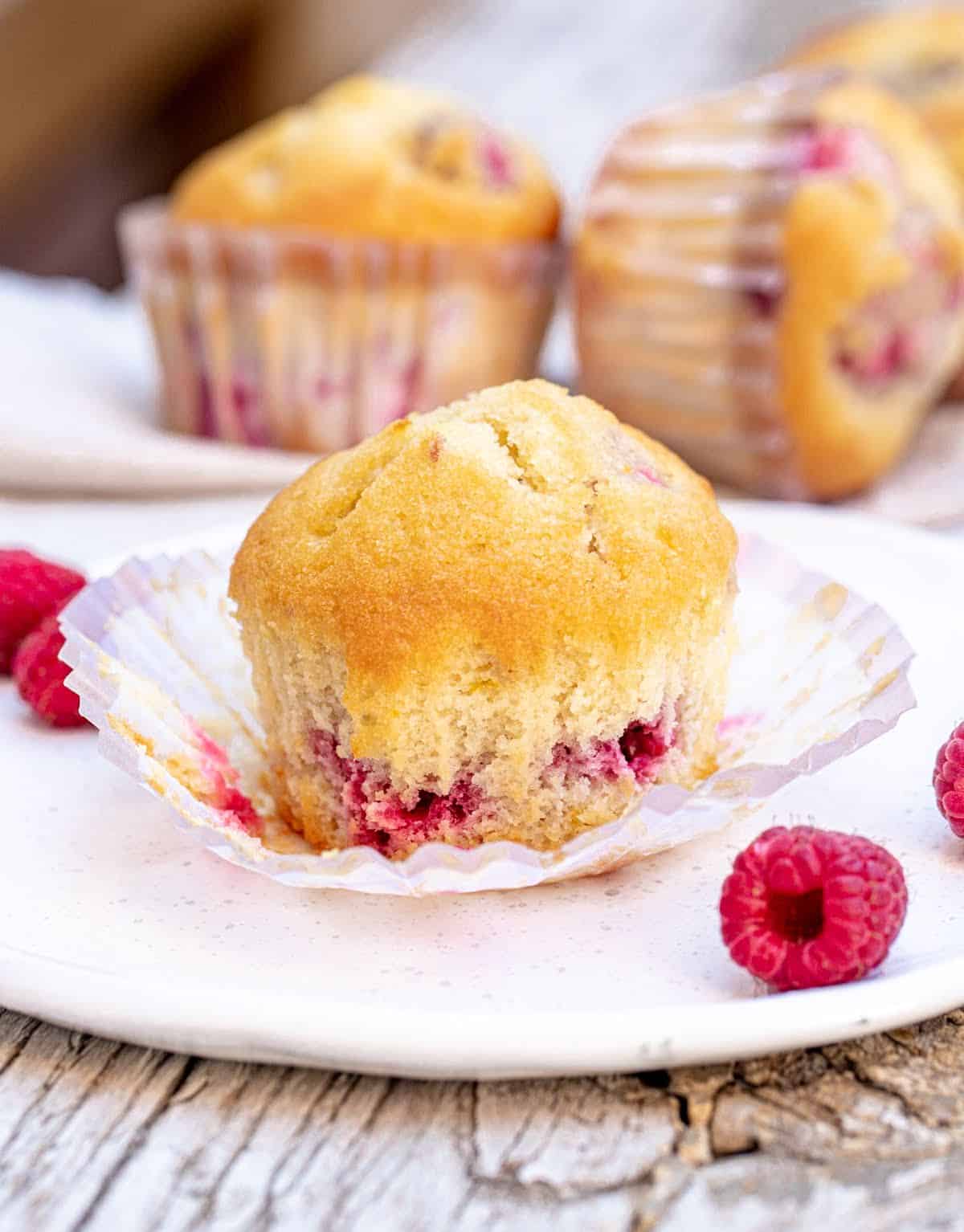 Whole raspberry muffin on opened paper liner on white plate, more muffins in background.