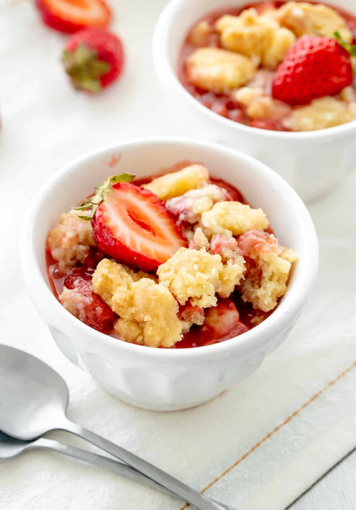 Two white bowls with servings of strawberry cobbler, white linen surface, a few fresh cut strawberries.