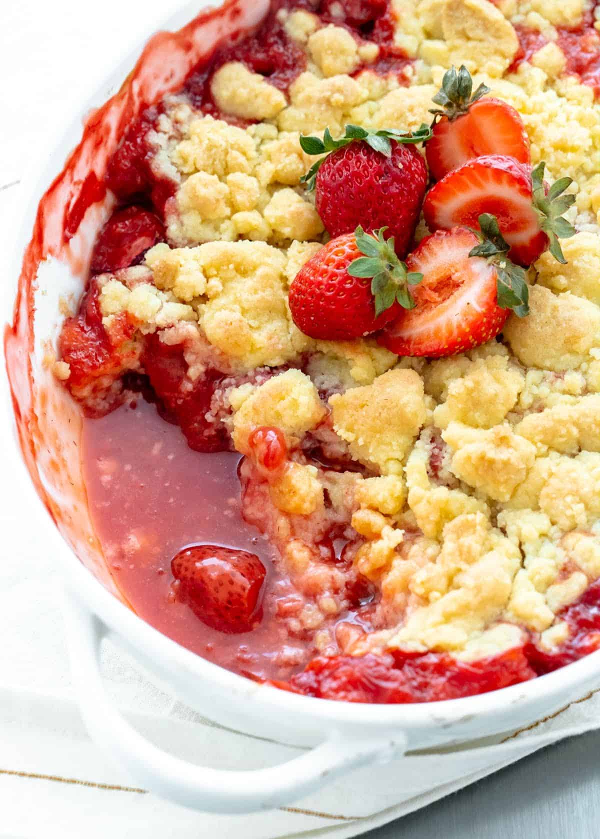 Partially eaten strawberry dump cake in a white ceramic dish with fresh strawberries on top. 