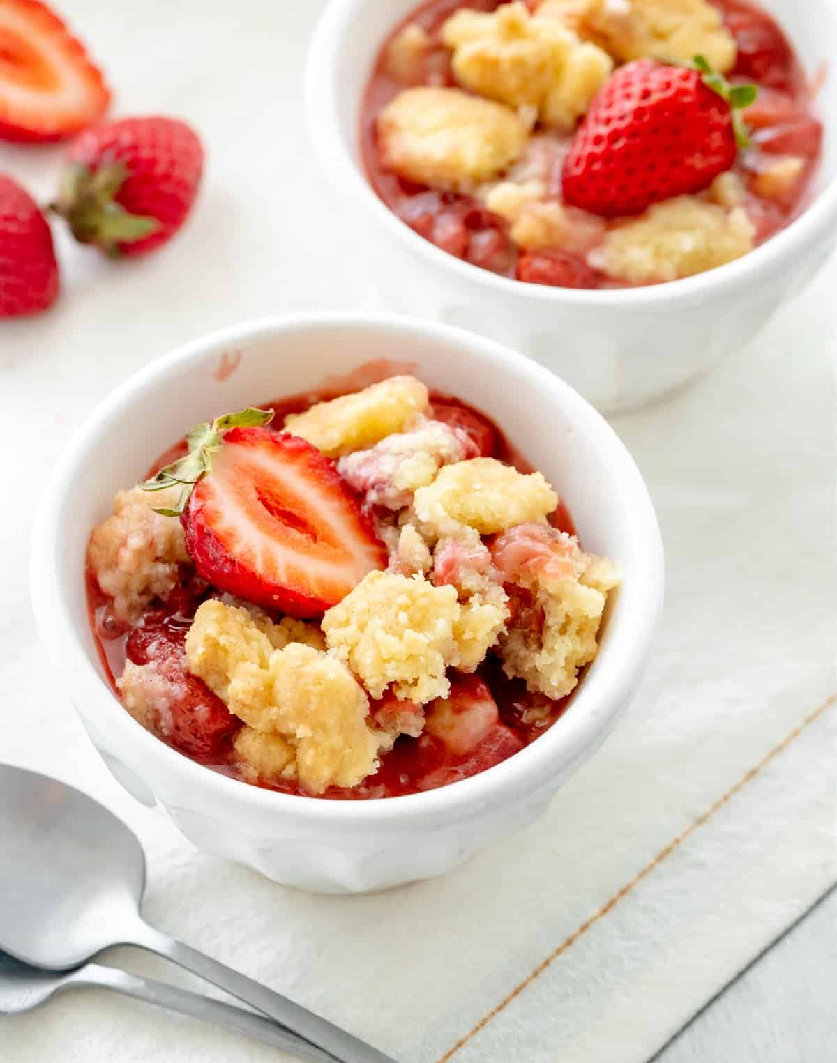 White bowls on white linen with servings of strawberry dump cake, fresh cut strawberries and silver spoon.