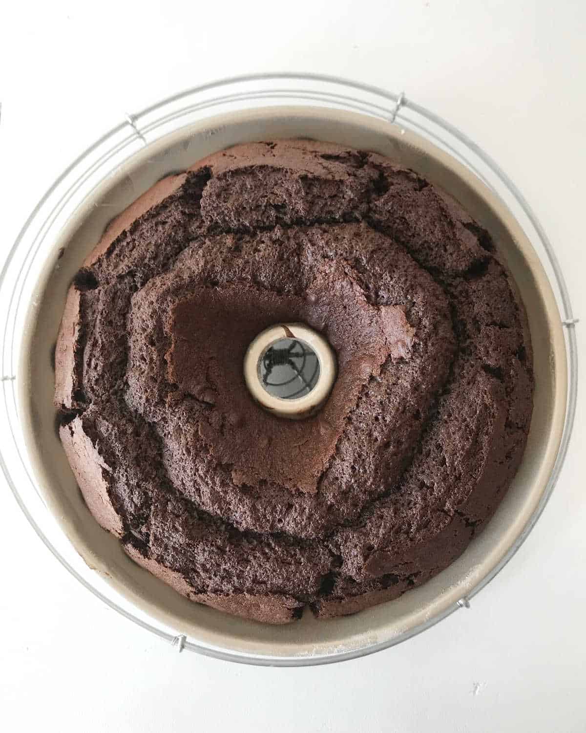 Baked chocolate bundt cake in the pan on a wire rack on a white surface. Top view.