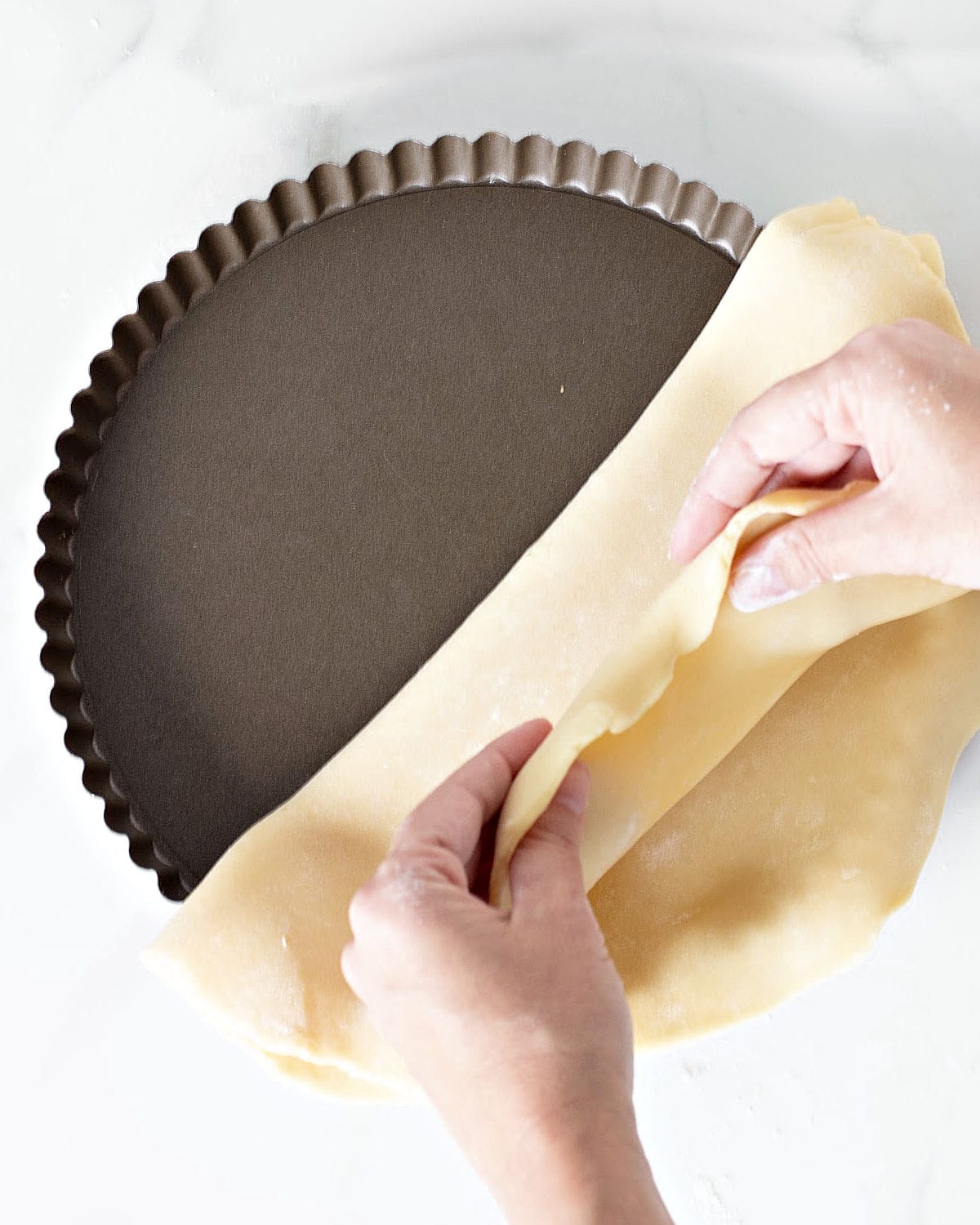 Hands lining a metal tart pan with pie crust. White surface. Top view.
