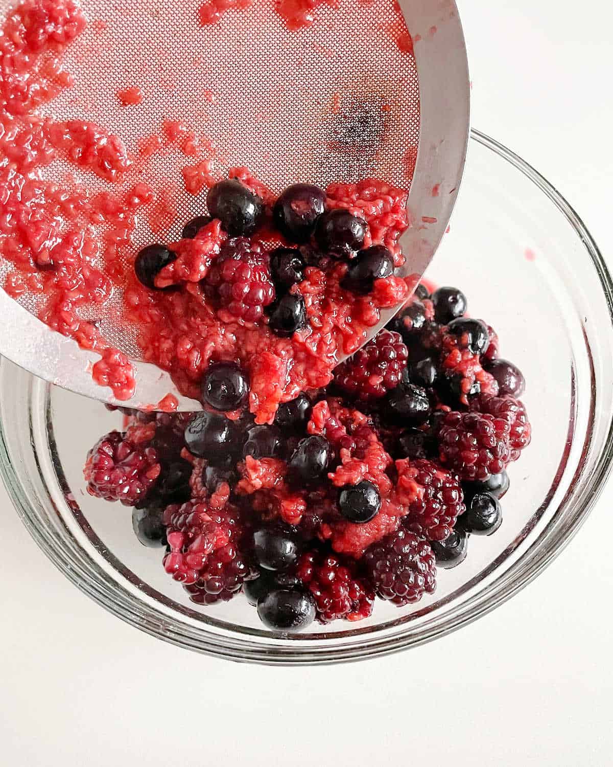 Straining cooked berries into a glass bowl on a white surface.