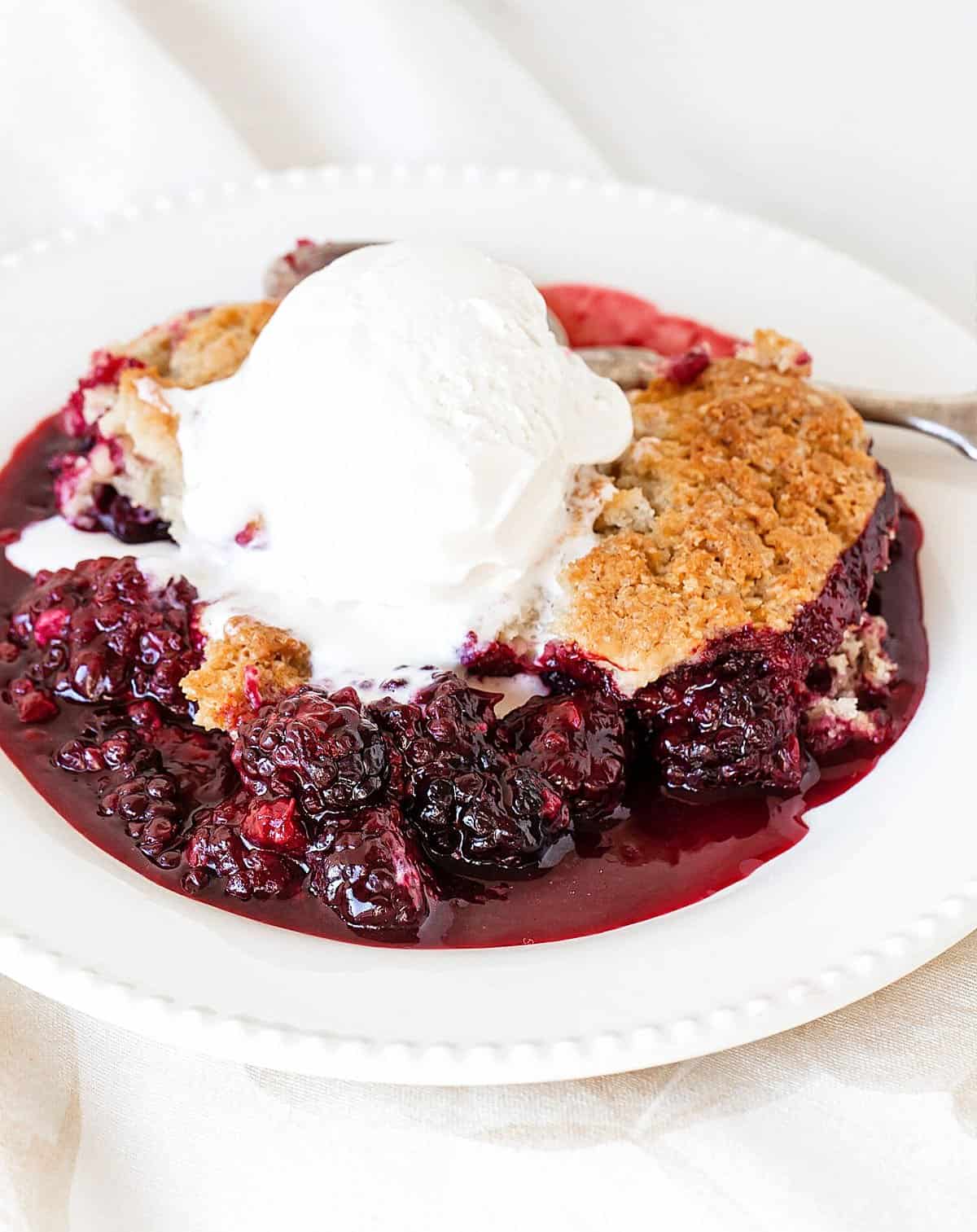 White plate with blackberry cobbler serving with ice cream, white background.