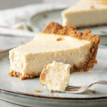 Single slice of cheesecake pie on grey plate, fork with bite. Another slice in background.