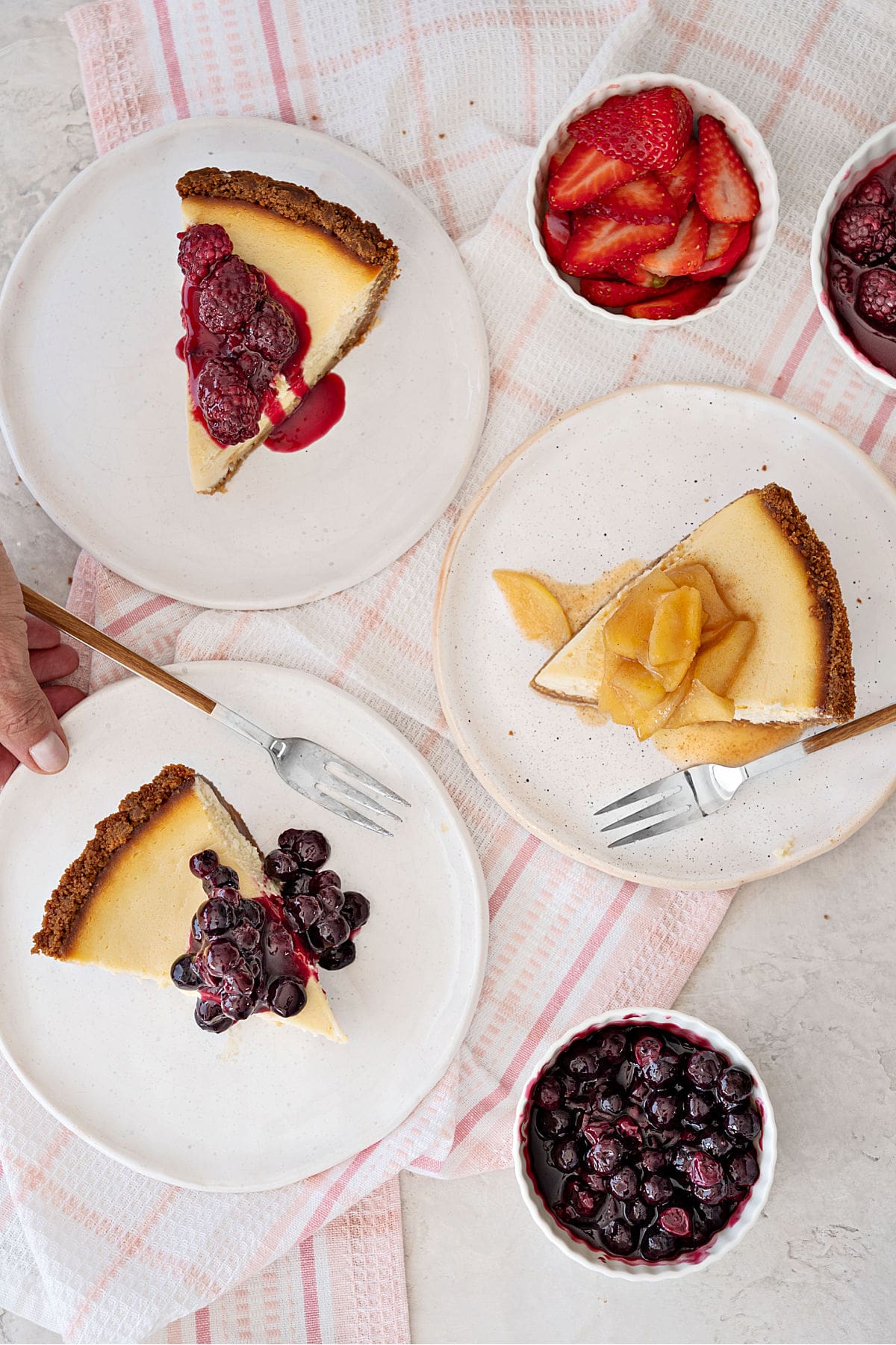 Slices of cheesecake pie on white plates with assorted fruit toppings. Pink striped kitchen towel. 