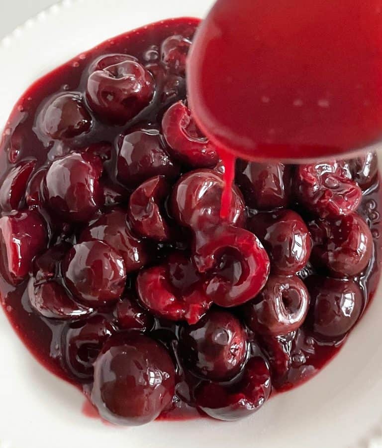 Syrup drizzling down from spoon onto cherry sauce in white plate.