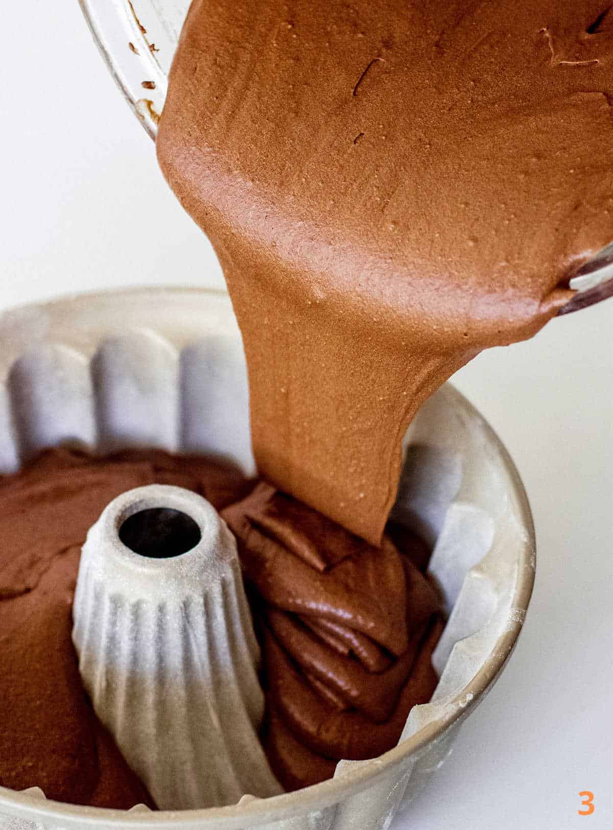 Pouring chocolate cake batter in metal bundt pan on white surface.
