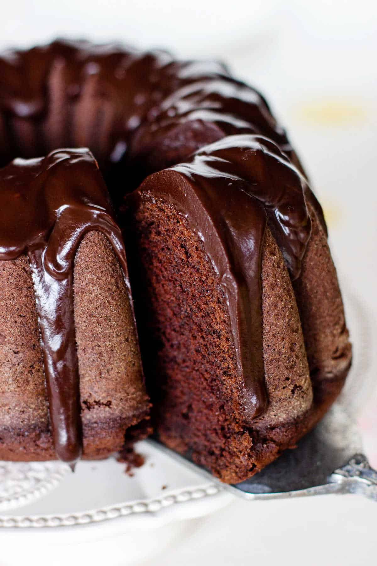 Close up of slice in cake server of glazed chocolate bundt cake on white plate.