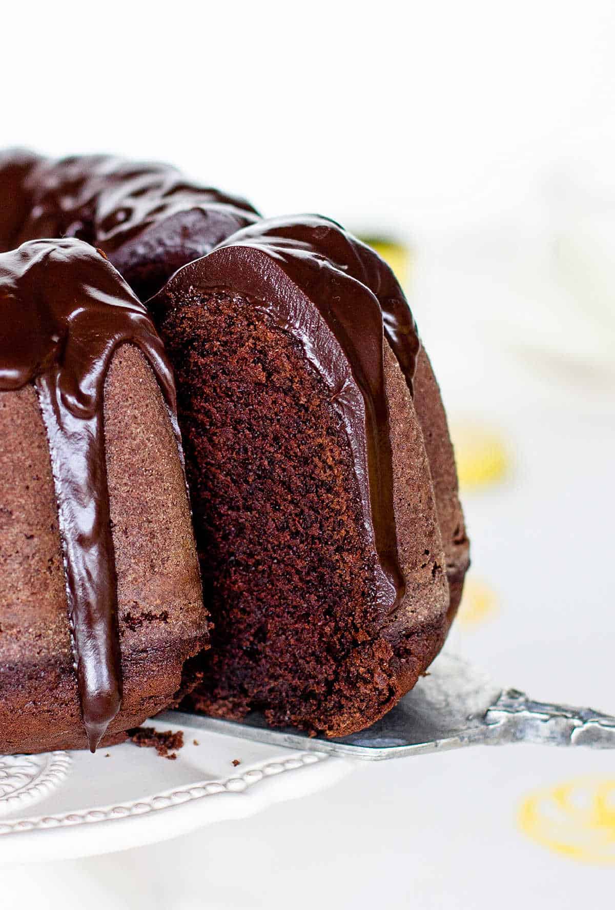 Removing slice from glazed chocolate bundt cake on white cake stand; white yellow background.