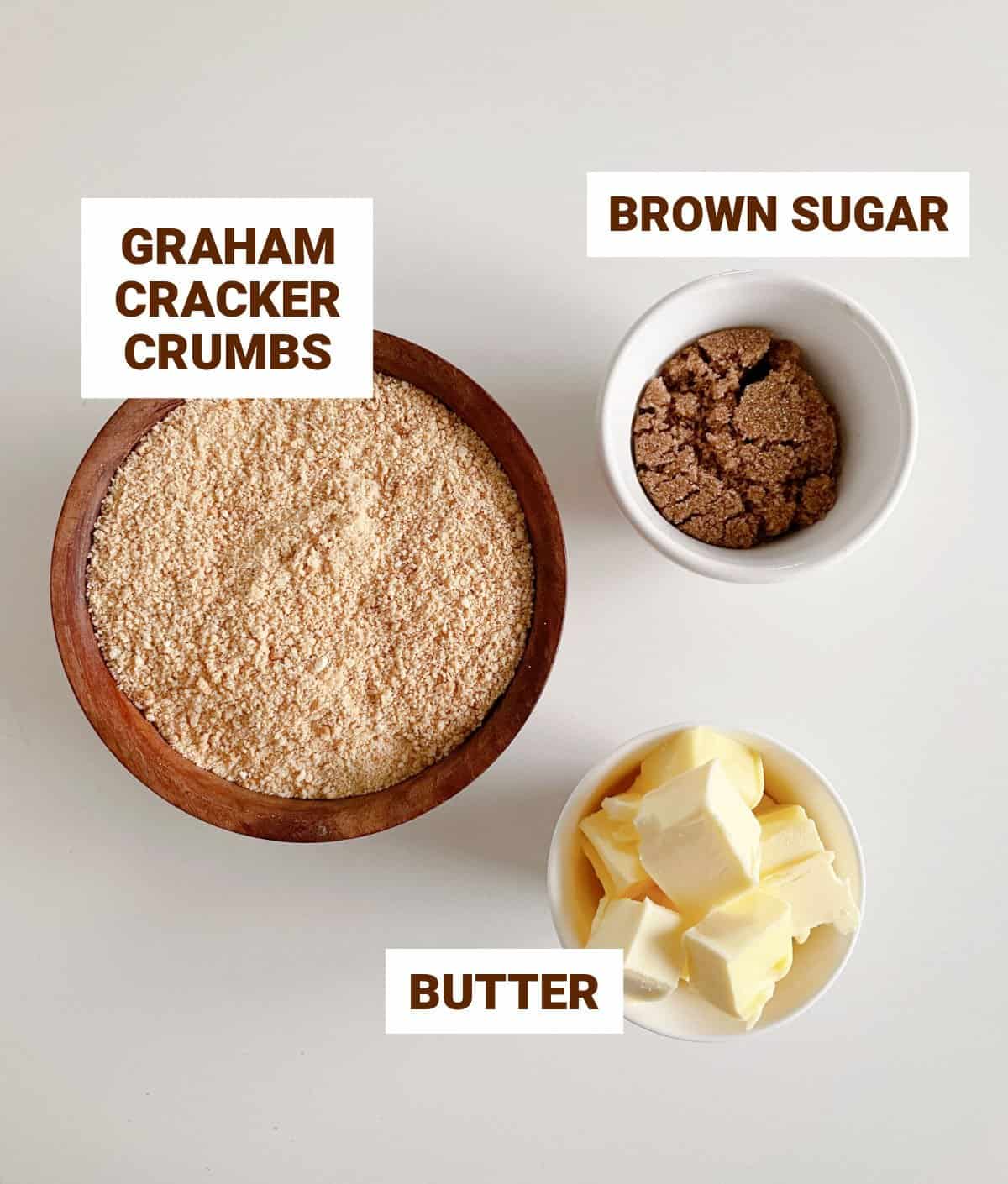 Light colored surface with bowls containing ingredients for graham cracker crust including brown sugar and butter.