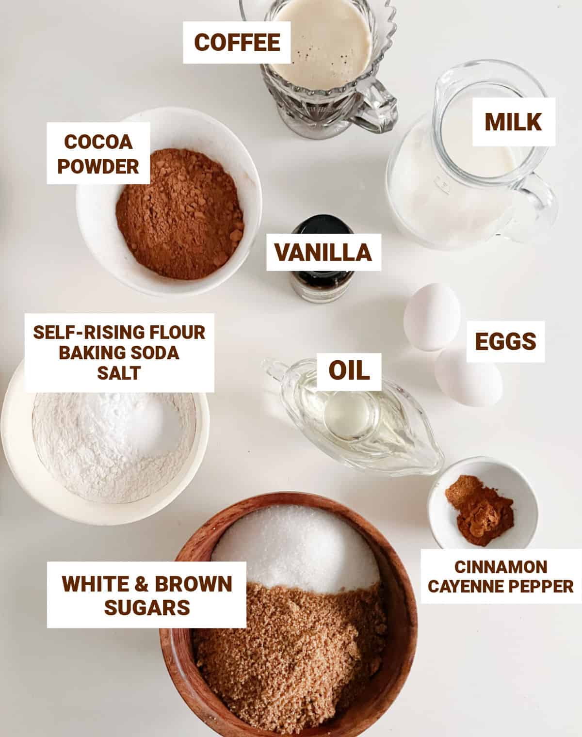 White surface with bowls containing ingredients for chocolate cake including cinnamon, spices, oil, coffee, milk, eggs, sugars, cocoa, vanilla, flour mixture.