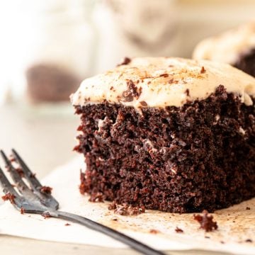 Close up square of chocolate cake with cinnamon frosting on beige parchment paper. A dark fork.