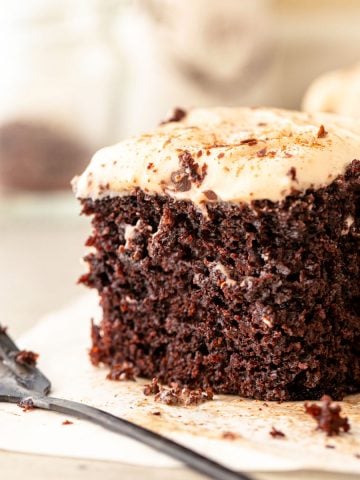 Close up square of chocolate cake with cinnamon frosting on beige parchment paper. A dark fork.