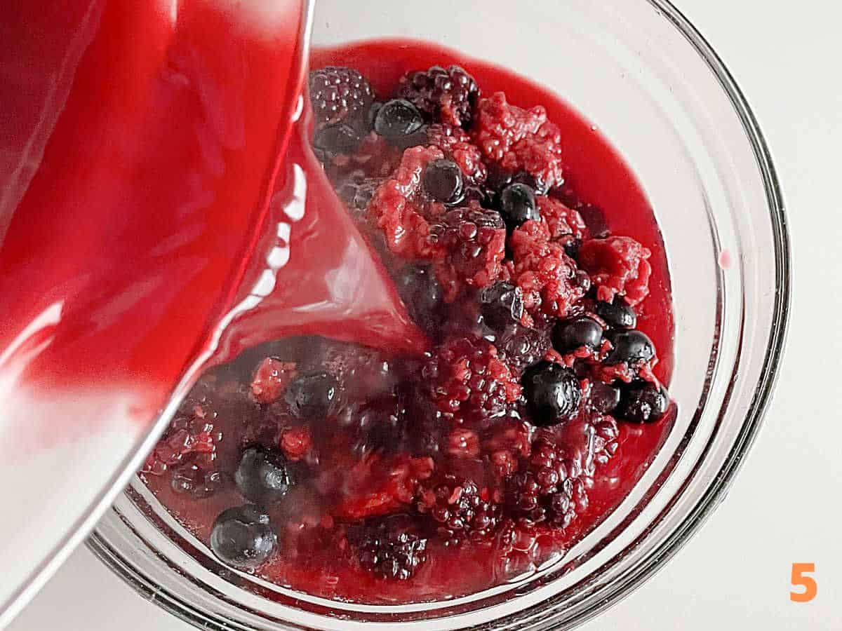 Red juice being poured over berries in glass bowl on a white surface.