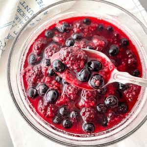 Close up top view of glass bowl with mixed berry compote, silver spoon above, white linen underneath.