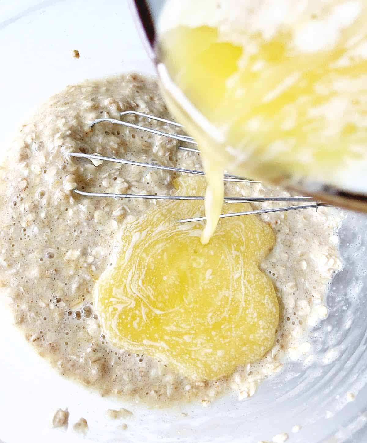 Close up of melted butter being poured over oatmeal mixture on a glass bowl with a whisk.