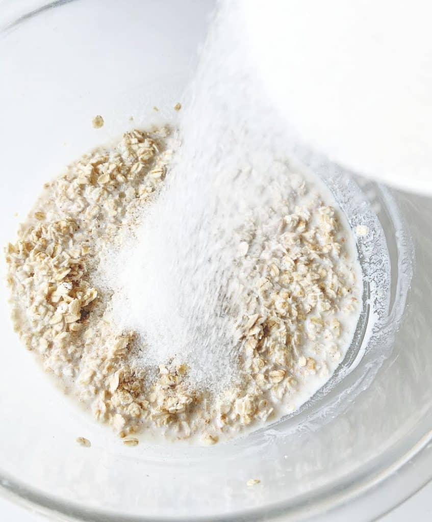 Sugar being added to oatmeal mixture in a glass bowl on a white surface.