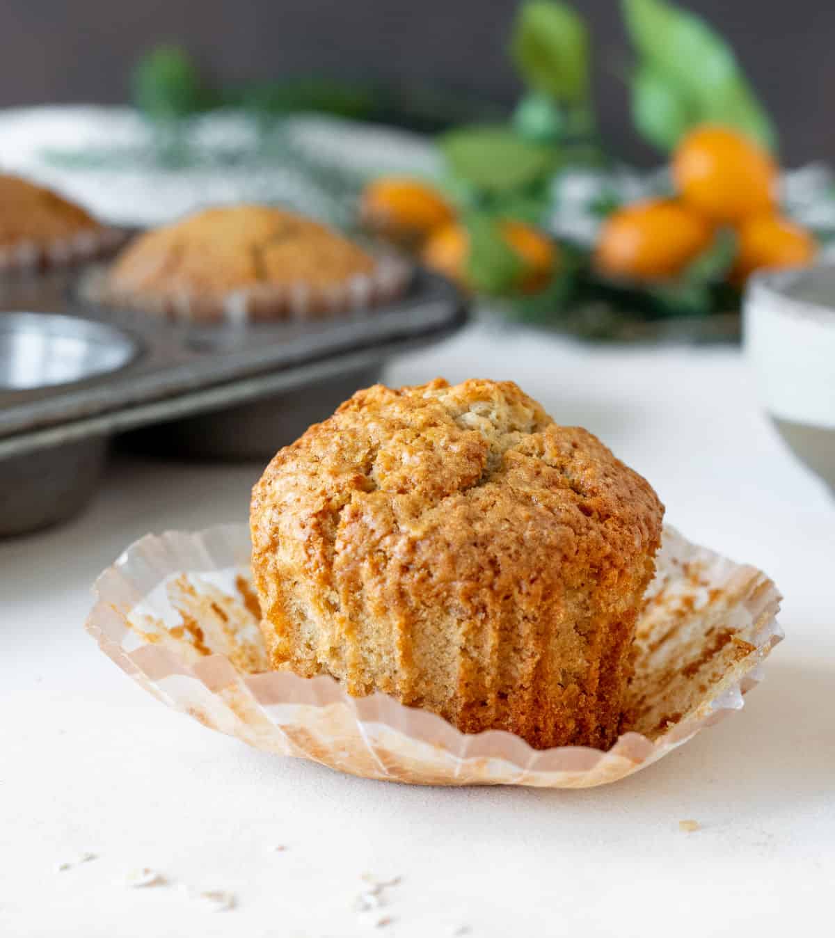 Single oatmeal muffin on an opened paper liner on a white surface. Muffin pan and kumquat branch in the background. 