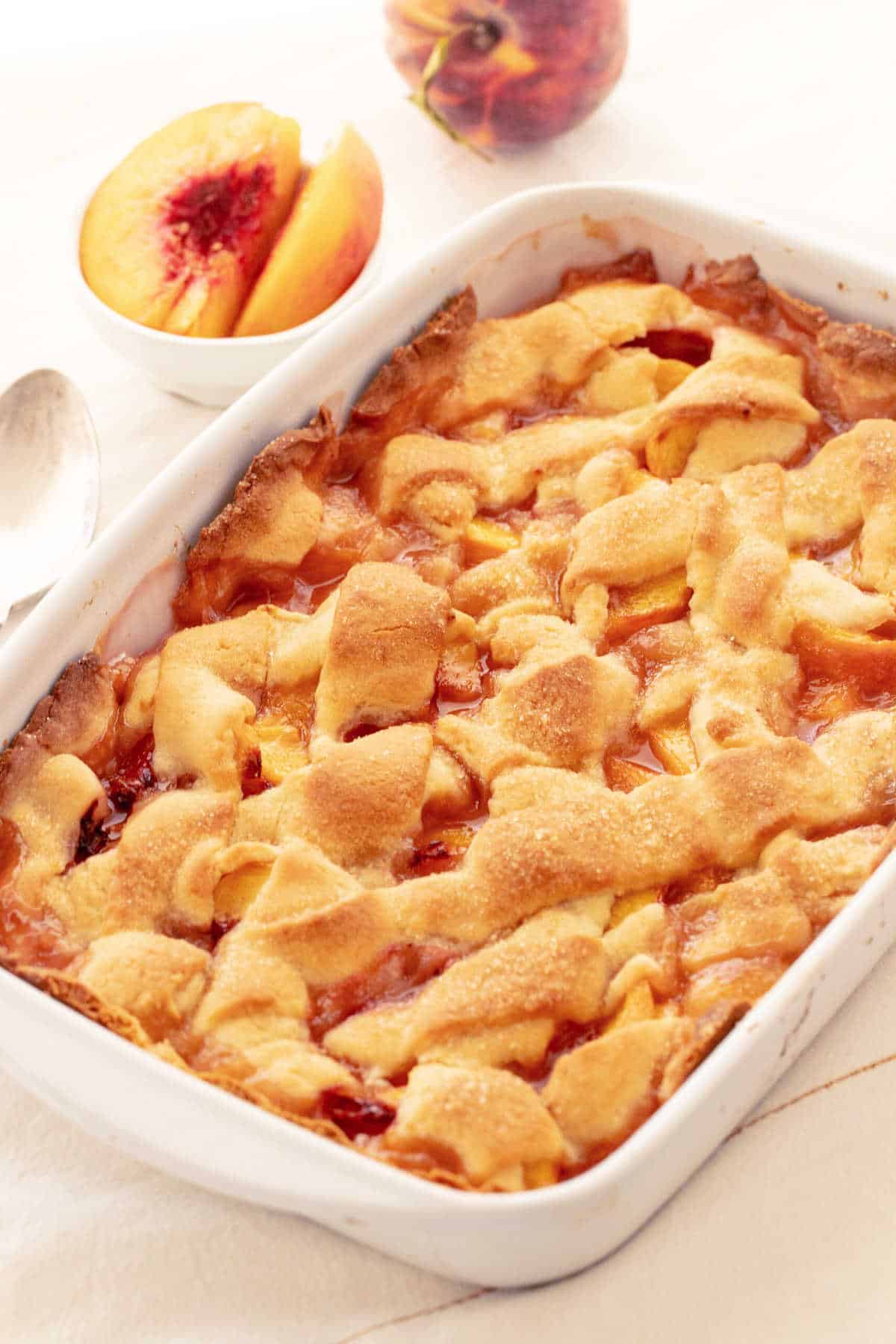 Rectangular dish with pie crust topped peach cobbler, white surface, fresh cut peaches in background.